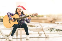 Jeune fille de métis jouant la guitare, chantant et souriant joyeux par la piscine, avec le chapeau de Santa de Noël image libre de droits Jeune fille de métis jouant la guitare, chantant et souriant joyeux par la piscine, avec le chapeau de Santa de Noël image libre de droits