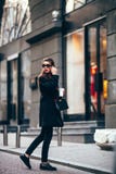 Jeune fille élégante, passant par Windows Verres à la mode de port et un manteau noir Conserve le café Photos libres de droits Jeune fille élégante, passant par Windows Verres à la mode de port et un manteau noir Conserve le café Photos libres de droits