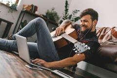 Jeune hippie de guitariste à la maison s'asseyant sur le plancher tenant l'ordinateur portable de lecture rapide de musique d'éco photo stock Jeune hippie de guitariste à la maison s'asseyant sur le plancher tenant l'ordinateur portable de lecture rapide de musique d'éco photo stock