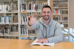 Jeune homme avec des pouces vers le haut d'heureux et du sourire avec un livre sur la table dans la bibliothèque images stock Jeune homme avec des pouces vers le haut d'heureux et du sourire avec un livre sur la table dans la bibliothèque images stock