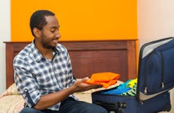 Jeune homme beau s'asseyant sur la valise d'emballage de lit et souriant, concept d'invité de pension image libre de droits Jeune homme beau s'asseyant sur la valise d'emballage de lit et souriant, concept d'invité de pension image libre de droits