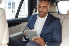 Jeune homme d'affaires s'asseyant sur le siège arrière dans la voiture passant en revue le comprimé numérique concentré photos stock Jeune homme d'affaires s'asseyant sur le siège arrière dans la voiture passant en revue le comprimé numérique concentré photos stock