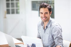 Jeune homme de sourire tenant des documents tout en se reposant au bureau avec l'ordinateur portable photographie stock libre de droits Jeune homme de sourire tenant des documents tout en se reposant au bureau avec l'ordinateur portable photographie stock libre de droits
