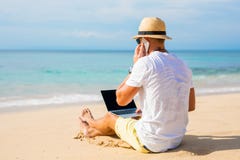 Jeune homme sur la plage fonctionnant avec l'ordinateur portable et parlant du téléphone image libre de droits Jeune homme sur la plage fonctionnant avec l'ordinateur portable et parlant du téléphone image libre de droits