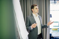 Jeune meneur d'équipe ou directeur sûr se tenant devant un tableau de conférence et un présent projet sur le bureau images stock Jeune meneur d'équipe ou directeur sûr se tenant devant un tableau de conférence et un présent projet sur le bureau images stock
