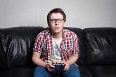 Jeune type avec les verres et la chemise rouge jouant des jeux vidéo sur la manette, se reposant sur un sofa en cuir noir image stock Jeune type avec les verres et la chemise rouge jouant des jeux vidéo sur la manette, se reposant sur un sofa en cuir noir image stock