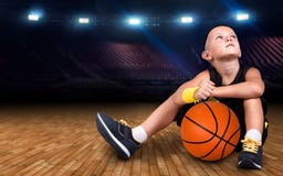 Jogador de basquetebol do menino com uma bola que senta-se no assoalho no gym e em sonhos de grandes vitórias fotografia de stock Jogador de basquetebol do menino com uma bola que senta-se no assoalho no gym e em sonhos de grandes vitórias fotografia de stock