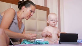 Jonge moeder die leuke baby met een lepel van havermoutpap voeden De babyzitting op het bed en staart beeldverhalen op tabletpc stock videobeelden Jonge moeder die leuke baby met een lepel van havermoutpap voeden De babyzitting op het bed en staart beeldverhalen op tabletpc stock videobeelden