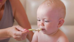 Jonge moeder die leuke baby met een lepel van havermoutpap voeden De babyzitting op het bed en staart beeldverhalen op tabletpc stock video Jonge moeder die leuke baby met een lepel van havermoutpap voeden De babyzitting op het bed en staart beeldverhalen op tabletpc stock video