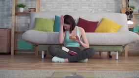 Jonge sportieve vrouwenzitting op de vloer die de badkamersschalen bekijken die een Hamburger eten stock video Jonge sportieve vrouwenzitting op de vloer die de badkamersschalen bekijken die een Hamburger eten stock video