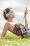 Jonge vrouw die aan muziek door MP3 speler luisteren die hoofdtelefoons met behulp van terwijl het liggen op gras tegen hemel Stock Afbeelding Jonge vrouw die aan muziek door MP3 speler luisteren die hoofdtelefoons met behulp van terwijl het liggen op gras tegen hemel Stock Afbeelding