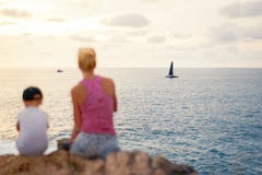 A jovem mulher com o menino que senta-se no penhasco na costa ensolarada perto da água e aprecia o por do sol fotografia de stock royalty free A jovem mulher com o menino que senta-se no penhasco na costa ensolarada perto da água e aprecia o por do sol fotografia de stock royalty free