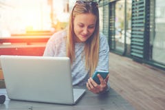 Jovem mulher com o smartphone e o portátil que sentam-se no café da rua, na Web da consultação e na alimentação social da rede foto de stock Jovem mulher com o smartphone e o portátil que sentam-se no café da rua, na Web da consultação e na alimentação social da rede foto de stock