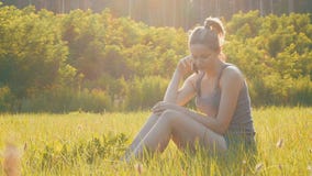 Jovem mulher feliz que senta-se no gramado verde e que fala no telefone ou no smartphone video estoque Jovem mulher feliz que senta-se no gramado verde e que fala no telefone ou no smartphone video estoque