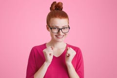 A jovem mulher freckled feliz do gengibre com nó do cabelo sorri felizmente, aperta os punhos como exulta exame com sucesso passa imagem de stock royalty free A jovem mulher freckled feliz do gengibre com nó do cabelo sorri felizmente, aperta os punhos como exulta exame com sucesso passa imagem de stock royalty free