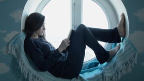 Jovem mulher que atravessa um livro de leitura A moça bonita no pijama senta-se em uma janela redonda que lê um livro filme Jovem mulher que atravessa um livro de leitura A moça bonita no pijama senta-se em uma janela redonda que lê um livro filme