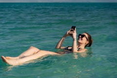 Jovem mulher que flutua na superfície da água do Mar Morto e que usa seu smartphone fotos de stock royalty free Jovem mulher que flutua na superfície da água do Mar Morto e que usa seu smartphone fotos de stock royalty free