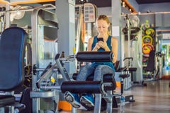 Jovem mulher que usa o telefone ao treinar no gym Mulher que senta-se em exercitar a m?quina que guarda o telefone celular imagens de stock royalty free Jovem mulher que usa o telefone ao treinar no gym Mulher que senta-se em exercitar a m?quina que guarda o telefone celular imagens de stock royalty free