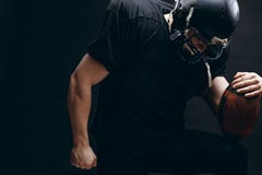 Jugador de fútbol americano con el casco y armadura con una bola contra la pared negra imagen de archivo libre de regalías Jugador de fútbol americano con el casco y armadura con una bola contra la pared negra imagen de archivo libre de regalías