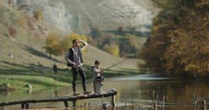 Jugando al muchacho con una caña de pescar grande, él está con su papá en la pesca al lado del lago almacen de metraje de vídeo Jugando al muchacho con una caña de pescar grande, él está con su papá en la pesca al lado del lago almacen de metraje de vídeo