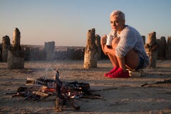 Junge Frau, die durch das Feuer an der Küste bei Sonnenuntergang sitzt lizenzfreies stockbild Junge Frau, die durch das Feuer an der Küste bei Sonnenuntergang sitzt lizenzfreies stockbild