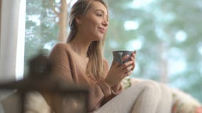 Junge glückliche Frau genießen von der Schale heißem Kaffee nach Hause sitzend durch das große Fenster mit Winterschnee-Baumhinte stock video Junge glückliche Frau genießen von der Schale heißem Kaffee nach Hause sitzend durch das große Fenster mit Winterschnee-Baumhinte stock video
