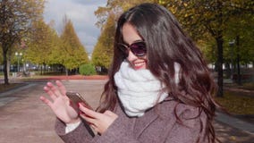 Junge lächelnde Brunettefrau mit den gekräuselten langen Haaren in der Sonnenbrille benutzt Smartphone im städtischen Park stock footage Junge lächelnde Brunettefrau mit den gekräuselten langen Haaren in der Sonnenbrille benutzt Smartphone im städtischen Park stock footage
