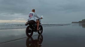 Junger h?bscher Hippie-Mann, der modernen kundenspezifischen Motorradrennl?ufer auf den schwarzen Sandstrand nahe dem Wasser reit stock footage Junger h?bscher Hippie-Mann, der modernen kundenspezifischen Motorradrennl?ufer auf den schwarzen Sandstrand nahe dem Wasser reit stock footage