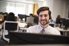 Junger Mann, der am Computer mit Kopfhörer im beschäftigten Büro arbeitet stockfotografie Junger Mann, der am Computer mit Kopfhörer im beschäftigten Büro arbeitet stockfotografie