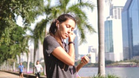Junges asiatisches Läufer-Mädchen setzt Kopfhörer an und dreht Musik am intelligenten Mobiltelefon im Stadt-Park 4K, Slowmotion stock video Junges asiatisches Läufer-Mädchen setzt Kopfhörer an und dreht Musik am intelligenten Mobiltelefon im Stadt-Park 4K, Slowmotion stock video