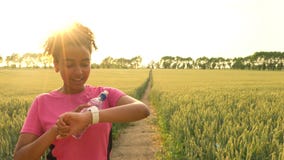 Mixed race African American girl teenager female young woman runner using smart watch and running on path through field at sunset