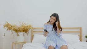 4K красивая азиатская женщина сидя на кровати слушая музыку с наушниками от применения на мобильном телефоне и танцевать видеоматериал 4K красивая азиатская женщина сидя на кровати слушая музыку с наушниками от применения на мобильном телефоне и танцевать видеоматериал