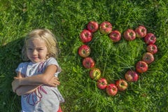 Kind blond jong Meisje met de rode vorm die van het appelenhart op het gras liggen stock afbeeldingen Kind blond jong Meisje met de rode vorm die van het appelenhart op het gras liggen stock afbeeldingen
