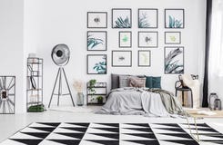 King-size bed with many pillows and grey sheets standing in whit. E bedroom interior with big studio lamp, black and white carpet and plastic chair with knit stock photos King-size bed with many pillows and grey sheets standing in whit. E bedroom interior with big studio lamp, black and white carpet and plastic chair with knit stock photos