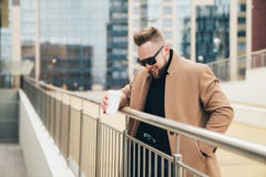 Knappe gebaarde jonge mens die in zonnebril kop van meeneemkoffie houden dichtbij commercieel centrum stock afbeeldingen Knappe gebaarde jonge mens die in zonnebril kop van meeneemkoffie houden dichtbij commercieel centrum stock afbeeldingen
