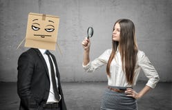 Kobieta jest przyglądająca przez magnifier przy biznesmenem z pudełkiem na jego głowie z pokerface zdjęcie royalty free Kobieta jest przyglądająca przez magnifier przy biznesmenem z pudełkiem na jego głowie z pokerface zdjęcie royalty free