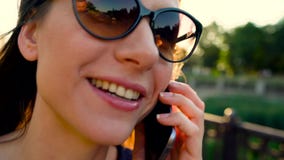 Kobieta opowiada na smartphone w okularach przeciwsłonecznych podczas gdy chodzący w dół ulicę przy zmierzchem, zakończenie up, z zbiory wideo Kobieta opowiada na smartphone w okularach przeciwsłonecznych podczas gdy chodzący w dół ulicę przy zmierzchem, zakończenie up, z zbiory wideo