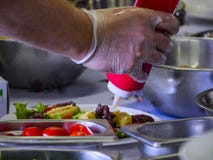 Kock Drizzling en sås från en röd flaska på en förberedd maträtt med rostfria bunkar omkring under att laga mat mästarklass, semi arkivbilder Kock Drizzling en sås från en röd flaska på en förberedd maträtt med rostfria bunkar omkring under att laga mat mästarklass, semi arkivbilder