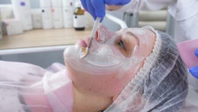 Kosmetiker setzt eine weiße Maske auf das Gesicht der Frau mit einer Bürste Hände eines Cosmetologist in den blauen Gummihandschu stock video Kosmetiker setzt eine weiße Maske auf das Gesicht der Frau mit einer Bürste Hände eines Cosmetologist in den blauen Gummihandschu stock video