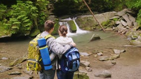 Kran geschossen: Ein paar Touristen mit Rucksäcken bewundern den schönen Wasserfall und den Gebirgsfluss Rückseitige Ansicht stock footage Kran geschossen: Ein paar Touristen mit Rucksäcken bewundern den schönen Wasserfall und den Gebirgsfluss Rückseitige Ansicht stock footage