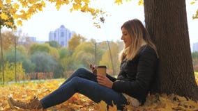 Kvinna som sitter nära träd i gula nedgångsidor, bruk Apps och dricker kaffe arkivfoton Kvinna som sitter nära träd i gula nedgångsidor, bruk Apps och dricker kaffe arkivfoton