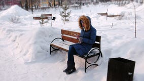 Kvinnan sitter på bänk, och bläddra mobiltelefonen i vinterstad parkera under dagen i snöig väder med att falla arkivfilmer Kvinnan sitter på bänk, och bläddra mobiltelefonen i vinterstad parkera under dagen i snöig väder med att falla arkivfilmer