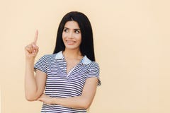 L'heureuse femelle de brune avec le sourire agréable, a de longs cheveux foncés, indique avec le doigt antérieur vers le haut, mo photo stock L'heureuse femelle de brune avec le sourire agréable, a de longs cheveux foncés, indique avec le doigt antérieur vers le haut, mo photo stock