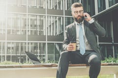 L'homme d'affaires dans le costume et le lien s'assied dehors sur le banc, café potable et parle à son téléphone portable Est tou Photographie stock L'homme d'affaires dans le costume et le lien s'assied dehors sur le banc, café potable et parle à son téléphone portable Est tou Photographie stock