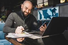 L'homme d'affaires travaille sur l'ordinateur portable, faisant des notes dans le document, parle dans des ?couteurs au t?l?phone image stock L'homme d'affaires travaille sur l'ordinateur portable, faisant des notes dans le document, parle dans des ?couteurs au t?l?phone image stock
