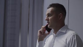 L'homme mignon de brune dans la chemise blanche parle du smartphone près de la fenêtre dans le bureau banque de vidéos L'homme mignon de brune dans la chemise blanche parle du smartphone près de la fenêtre dans le bureau banque de vidéos