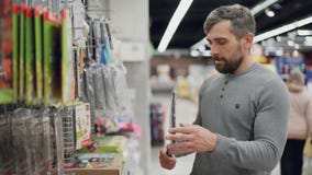 L'uomo barbuto bello sta scegliendo la griglia della griglia in supermercato, sta tenendolo che controlla la qualità ed il prezzo stock footage L'uomo barbuto bello sta scegliendo la griglia della griglia in supermercato, sta tenendolo che controlla la qualità ed il prezzo stock footage