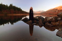 La belle dame avec les cheveux rouges, utilisant un manteau noir, s'est reflétée dans les eaux immobiles d'un lac Photographie stock libre de droits La belle dame avec les cheveux rouges, utilisant un manteau noir, s'est reflétée dans les eaux immobiles d'un lac Photographie stock libre de droits