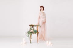 La belle femme enceinte avec les cheveux rouges se tient prêt le mur blanc avec la chaise décorée image stock La belle femme enceinte avec les cheveux rouges se tient prêt le mur blanc avec la chaise décorée image stock