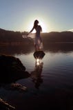 La belle fille s'est habillée dans le blanc, silhouetté par le soleil reflété dans le lac immobile Photo stock La belle fille s'est habillée dans le blanc, silhouetté par le soleil reflété dans le lac immobile Photo stock